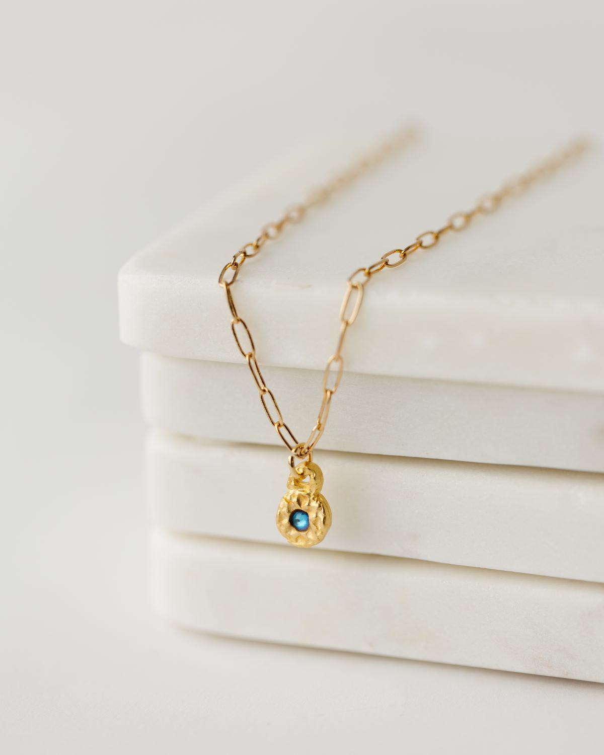 A gold-plated necklace with a small pendant featuring an azure CZ stone inlay, displayed on a white marble surface.