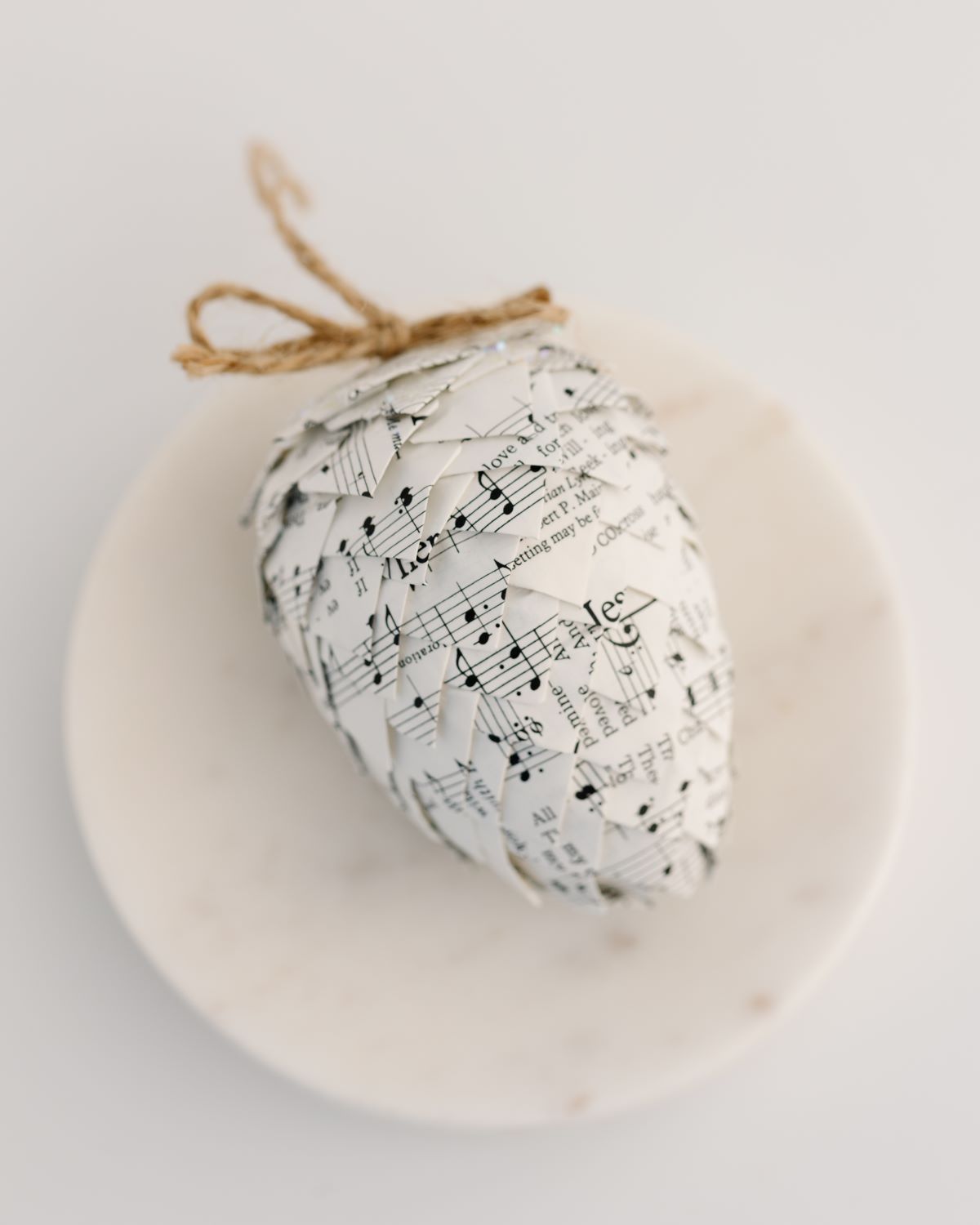 A pinecone ornament made from folded paper with a musical score design, tied with a string to a marble slice.