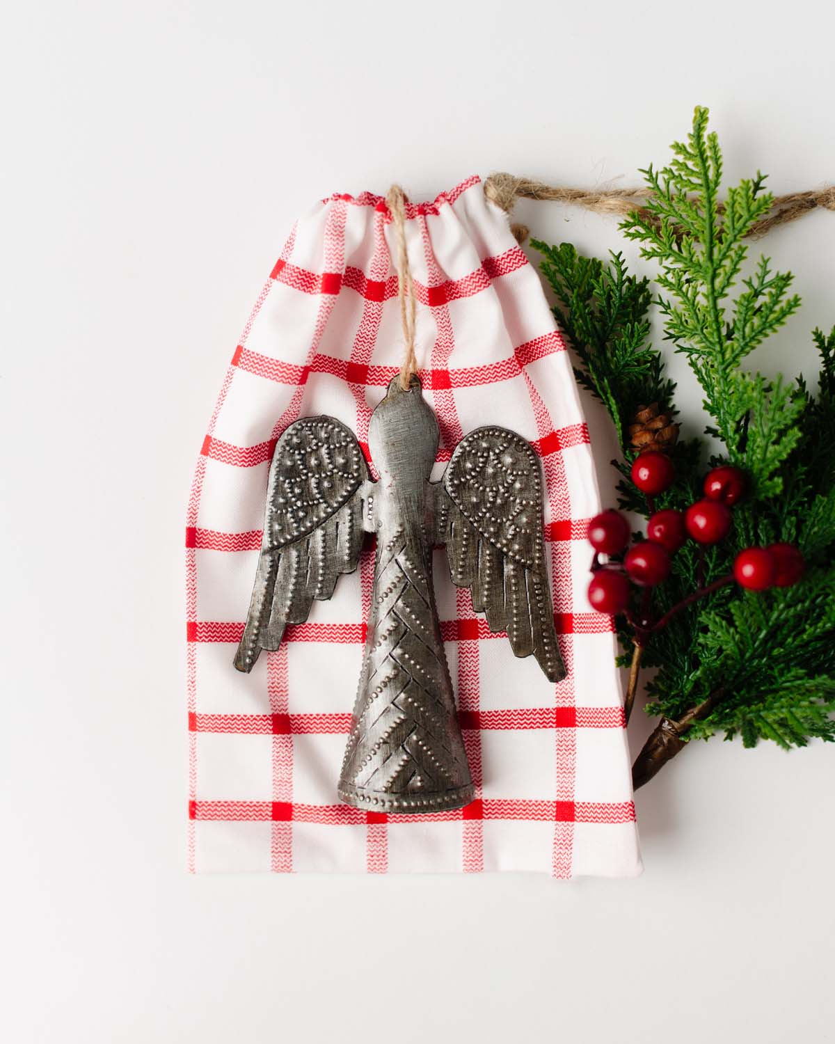 A metal angel ornament hanging from a red and white gingham fabric gift bag, with greenery and a red checkered pattern in the background.