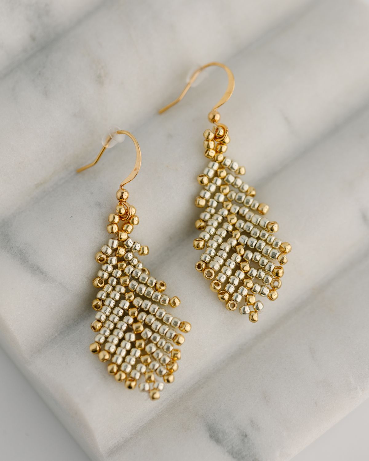 A pair of handcrafted gold and brass glass seed bead earrings laid on a marble surface.