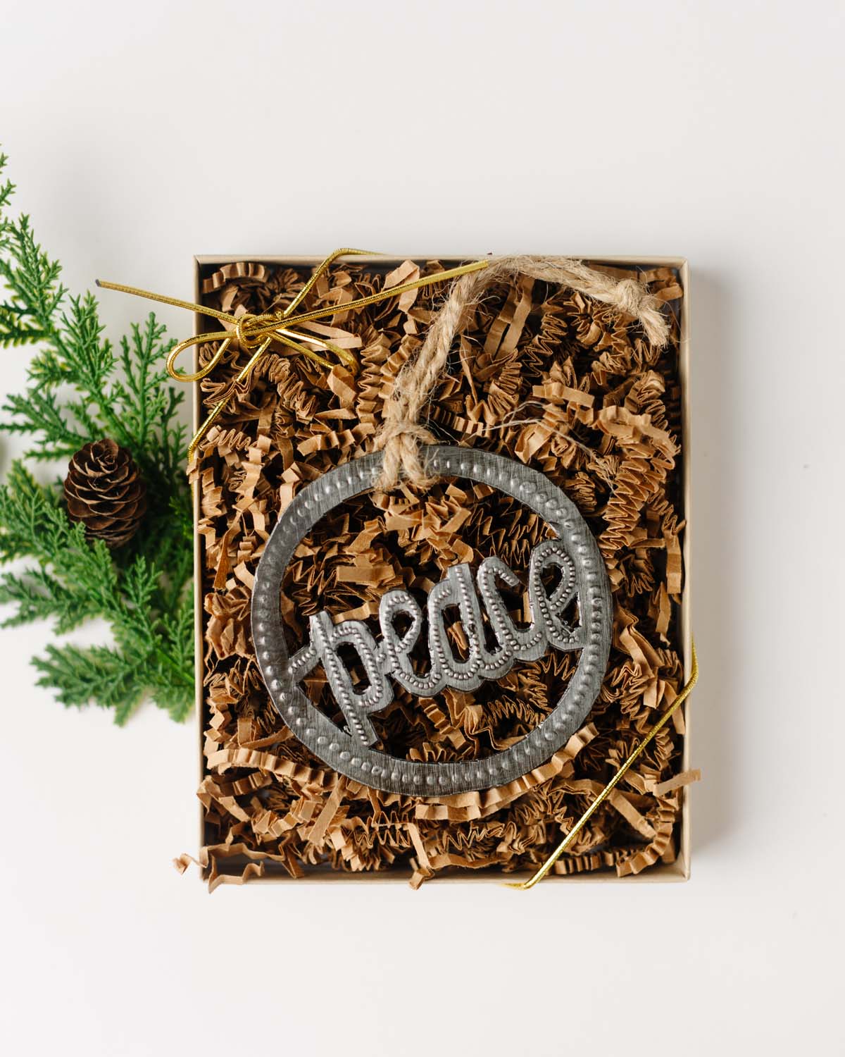 A metal ornament with the word 'peace' written on it, placed inside a kraft paper box with a clear lid, adorned with brown krinkle paper and a gold ribbon.