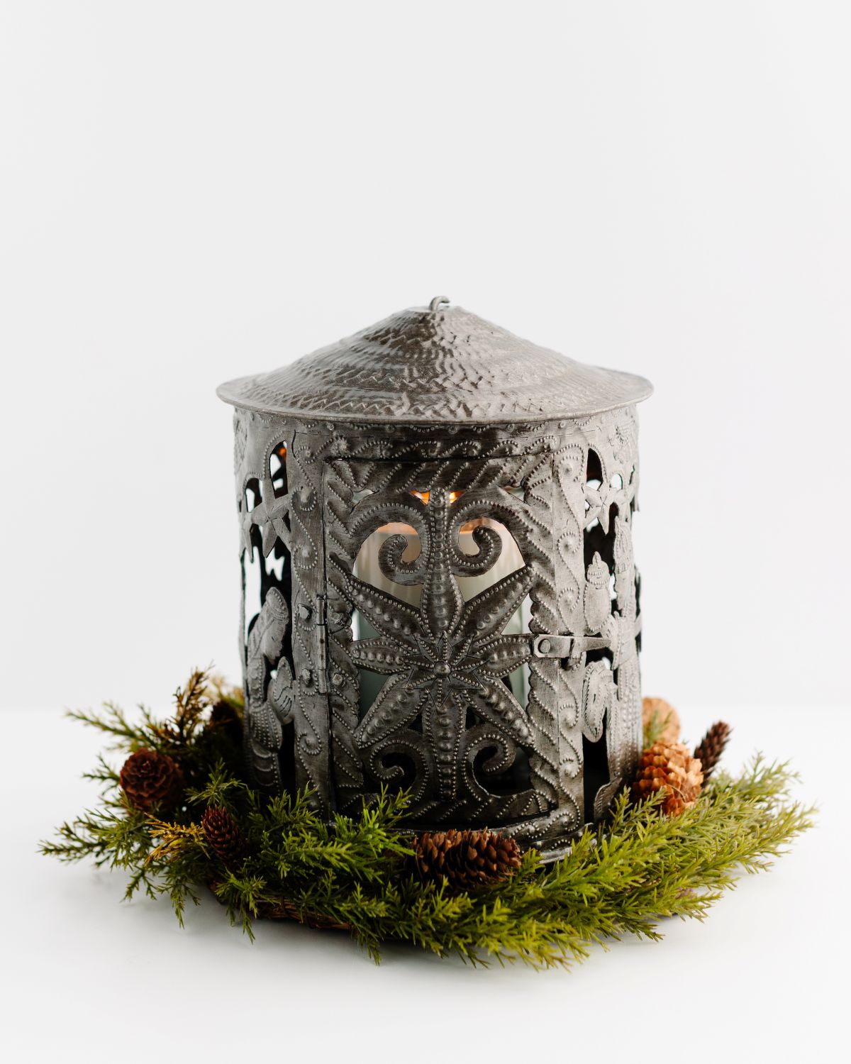 Decorative metal lantern with intricate patterns, surrounded by greenery and pinecones on a white background