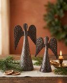 Two dark metal angels with textured wings and bell-shaped bodies, styled on a wooden table with pine branches, dried orange slices, and a lit candle in a brass holder against a warm brown background.